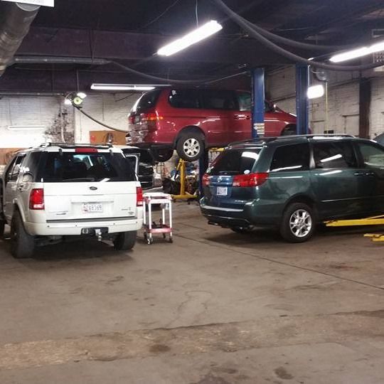 An indoor auto repair shop with a red van on a lift, a white SUV, and a dark green minivan parked on the concrete floor.
