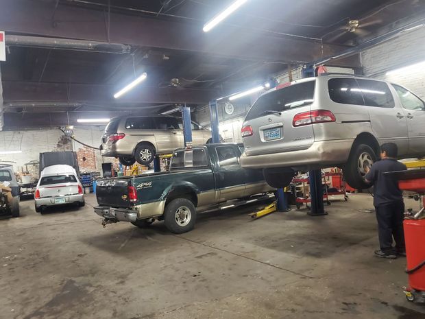 A mechanic works in a garage with several vehicles, including a van lifted on a hoist and a truck parked in the bay.