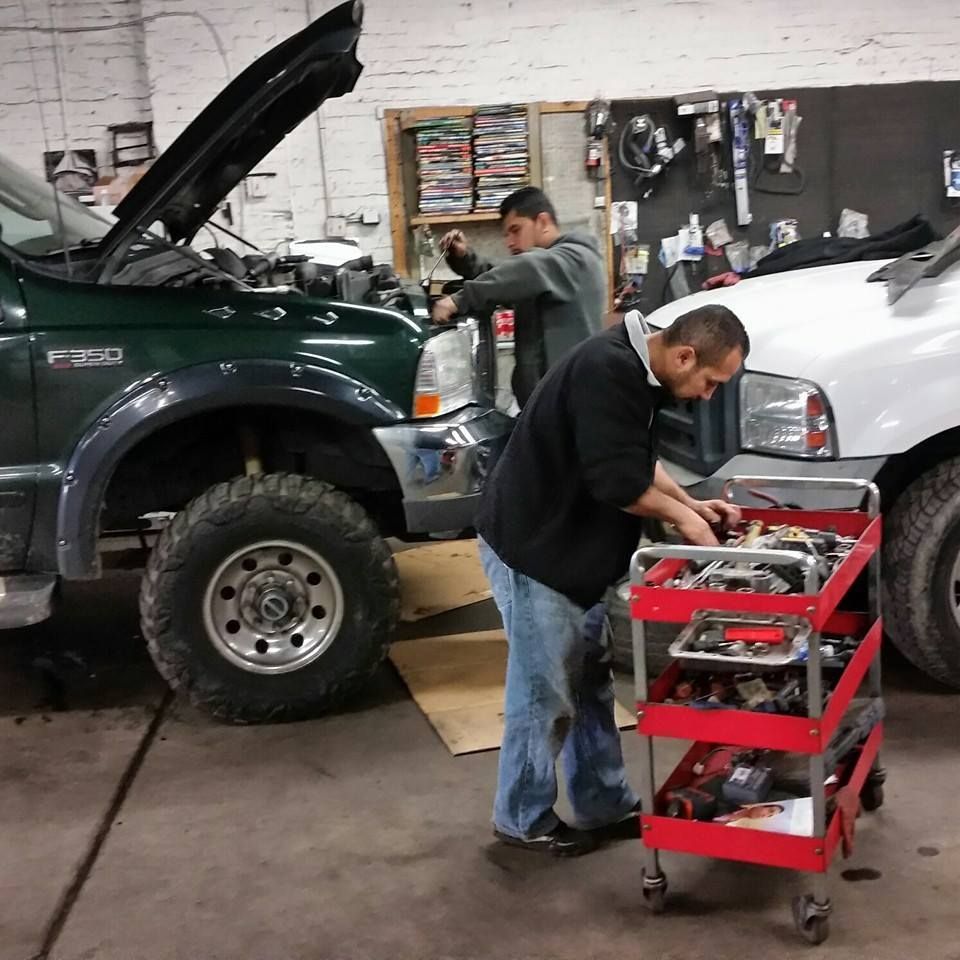 Two mechanics work on vehicles inside a repair shop, with one person selecting tools from a red rolling cart.