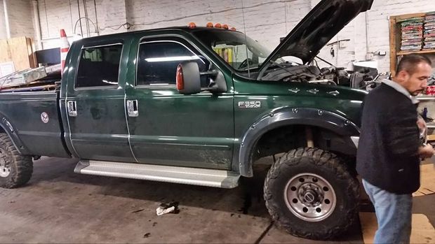 A mechanic works on the engine of a dark green Ford F-350 pickup truck with its hood open inside a garage.