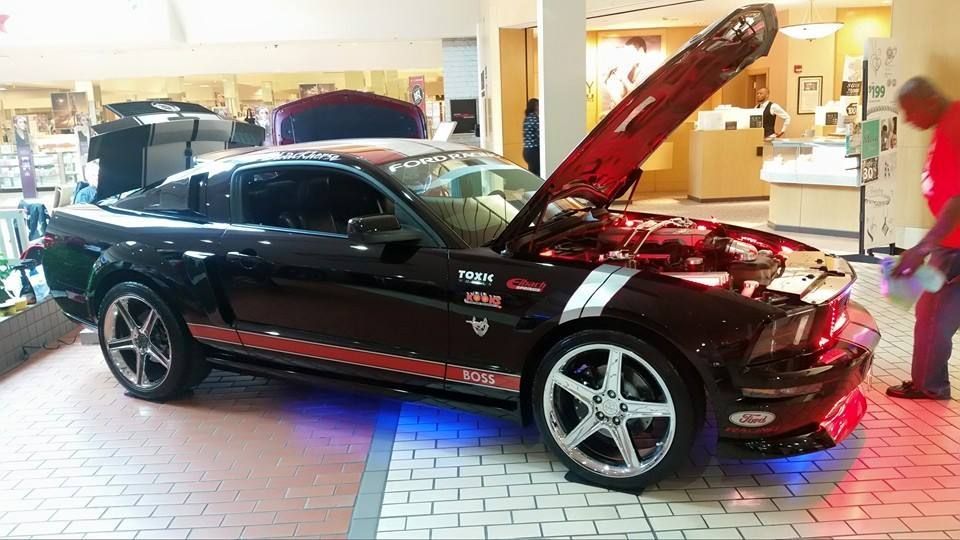 A black Ford Mustang with red racing stripes and blue underglow, displayed indoors with its hood open for a car show.