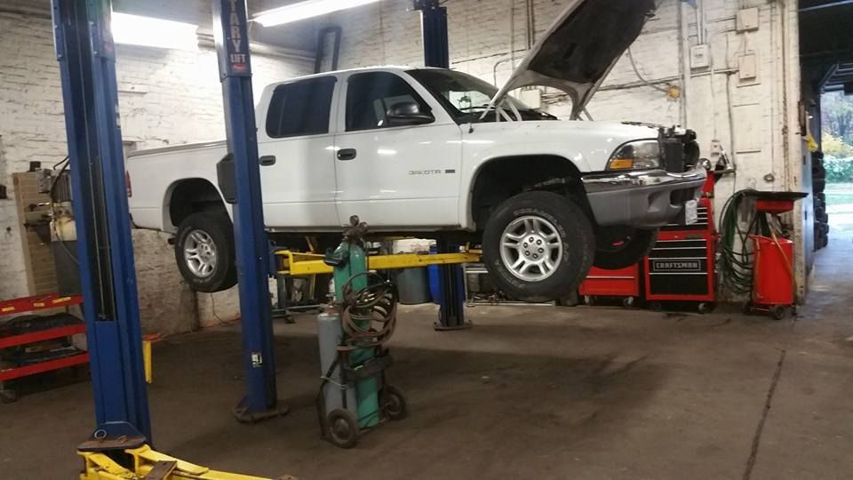 A white pickup truck with its hood open, raised on a hydraulic lift inside an auto repair shop.