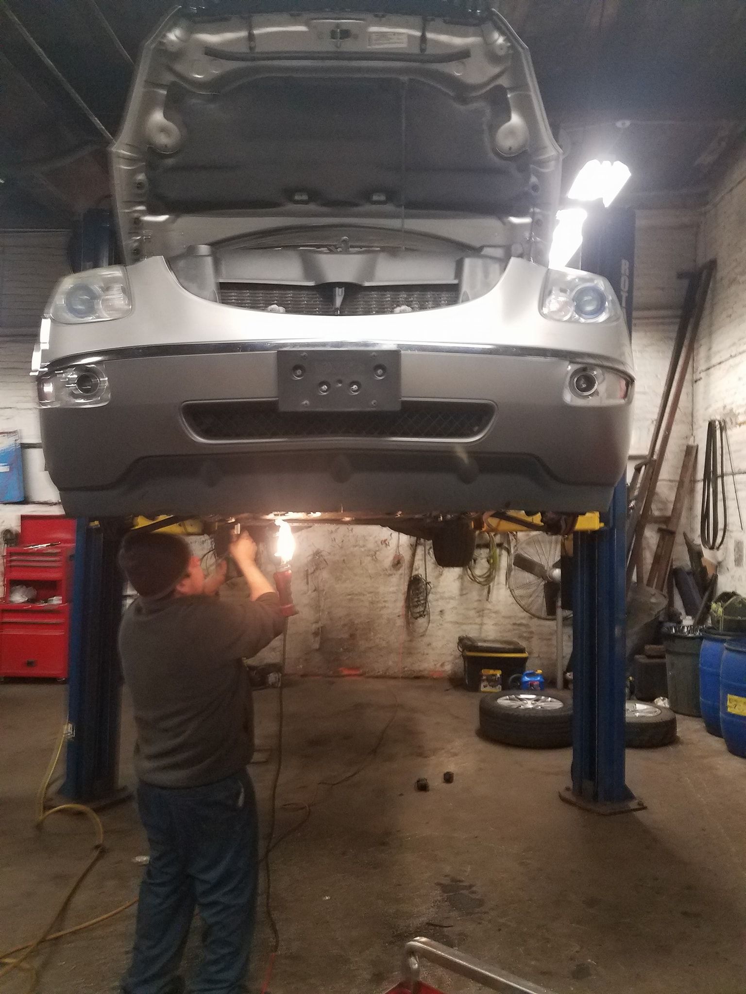 A person uses a welding tool on the undercarriage of a silver vehicle elevated on a lift inside an auto repair shop.