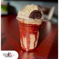 Red velvet milkshake in a clear cup, topped with whipped cream and an Oreo cookie, on a wooden table.