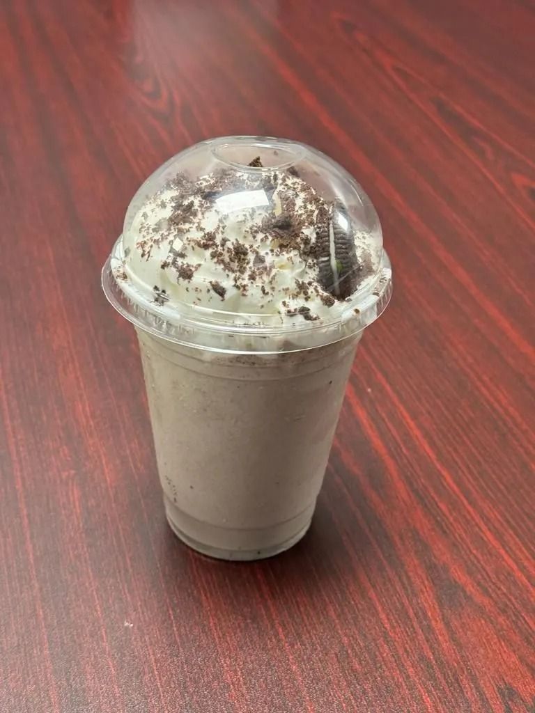 Oreo milkshake in a plastic cup, topped with whipped cream and cookie crumbs, on a wooden table.