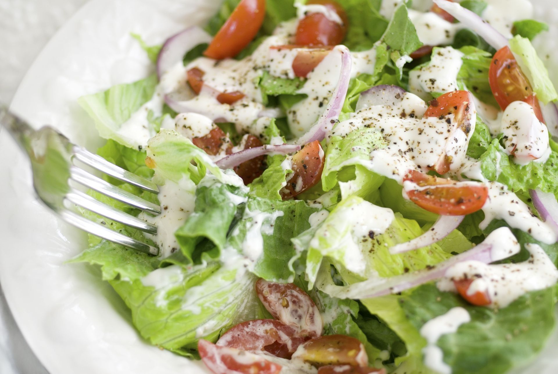 Salad with lettuce, tomatoes, red onion, and creamy dressing on a white plate; a fork is taking a bite.