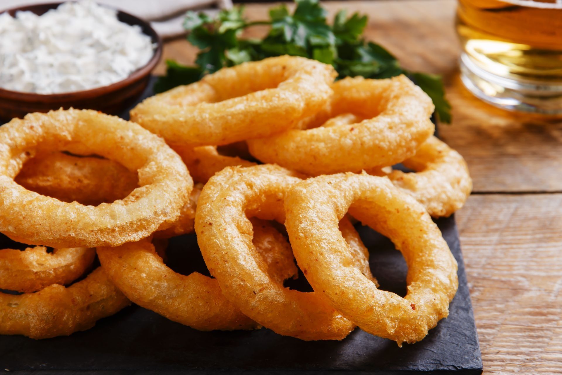 Golden fried onion rings piled on a black serving platter with a bowl of dipping sauce and a beer.