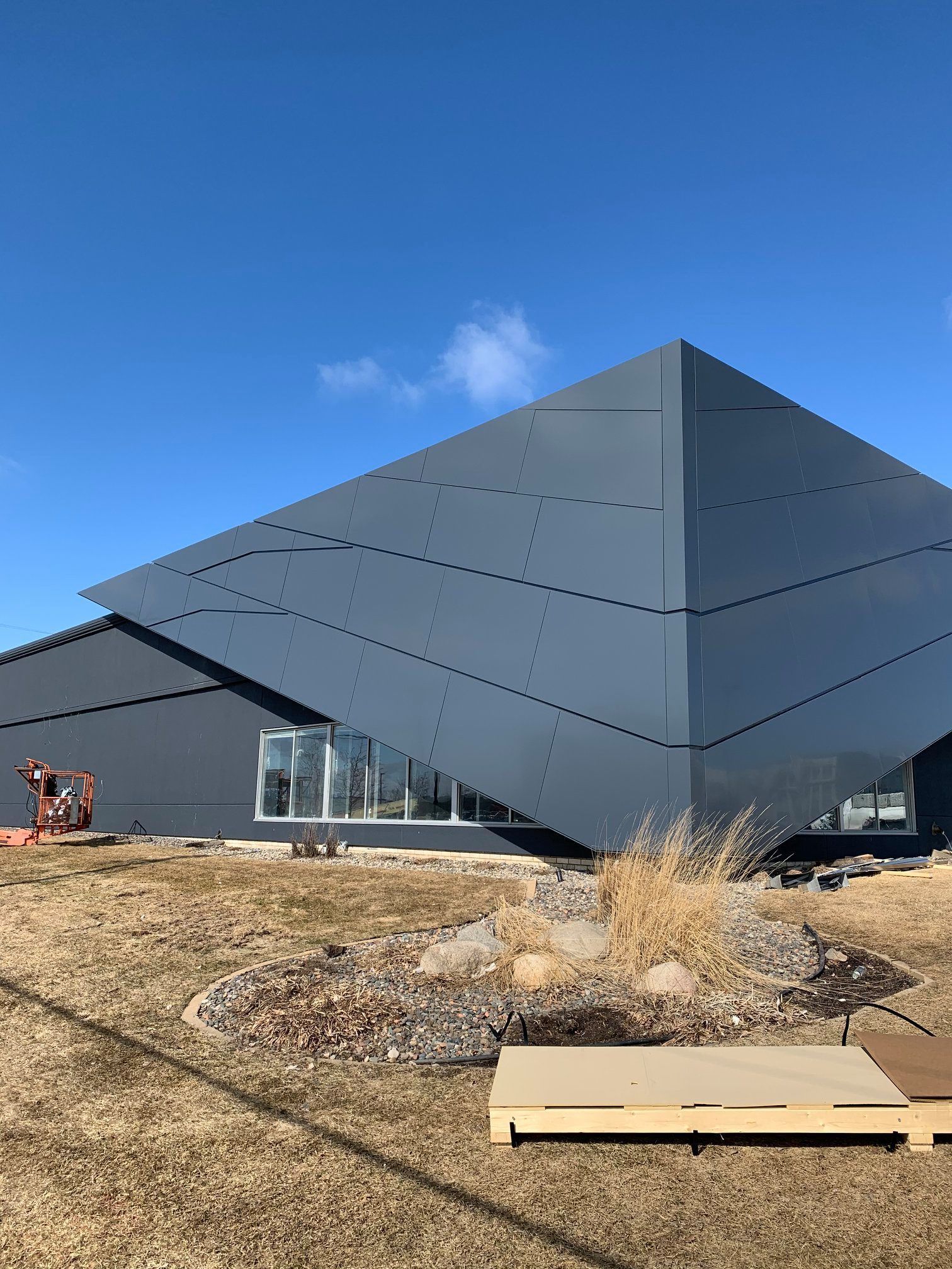 Modern gray building with angular design against a blue sky, dry grass in foreground.