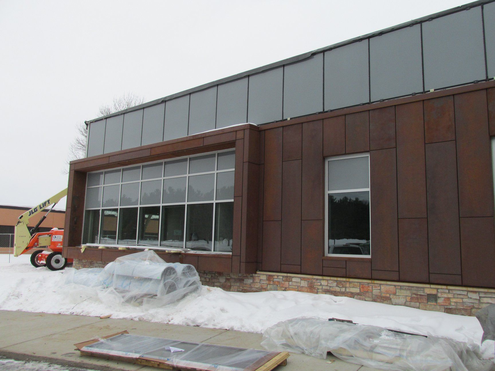 Building exterior with large windows, brown siding, and snow on the ground; lift in the left corner.