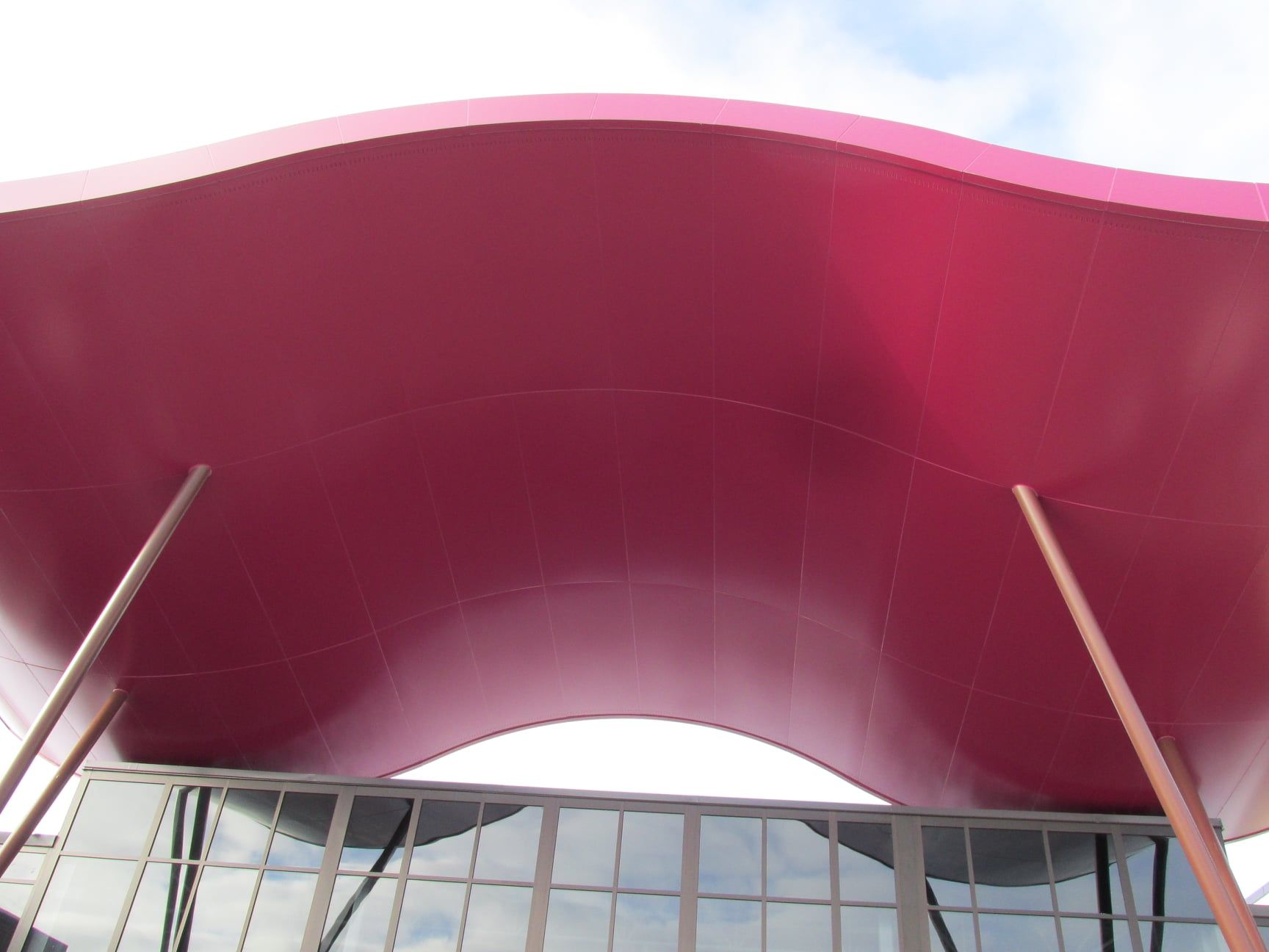 Pink wavy awning supported by two brown poles above a building with glass windows.