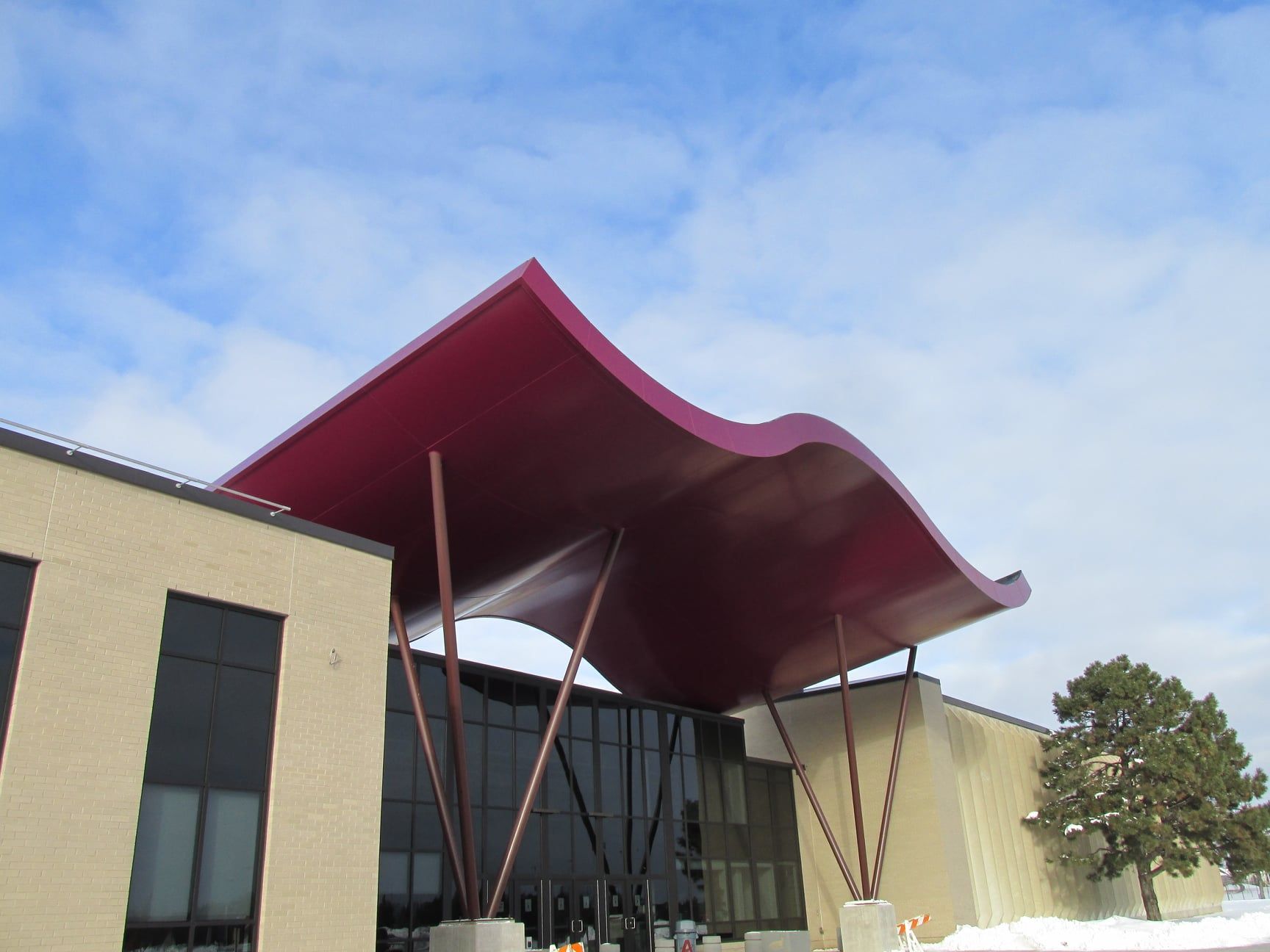 Building entrance with a wavy, magenta roof supported by bronze columns. Sunny day, cloudy sky.