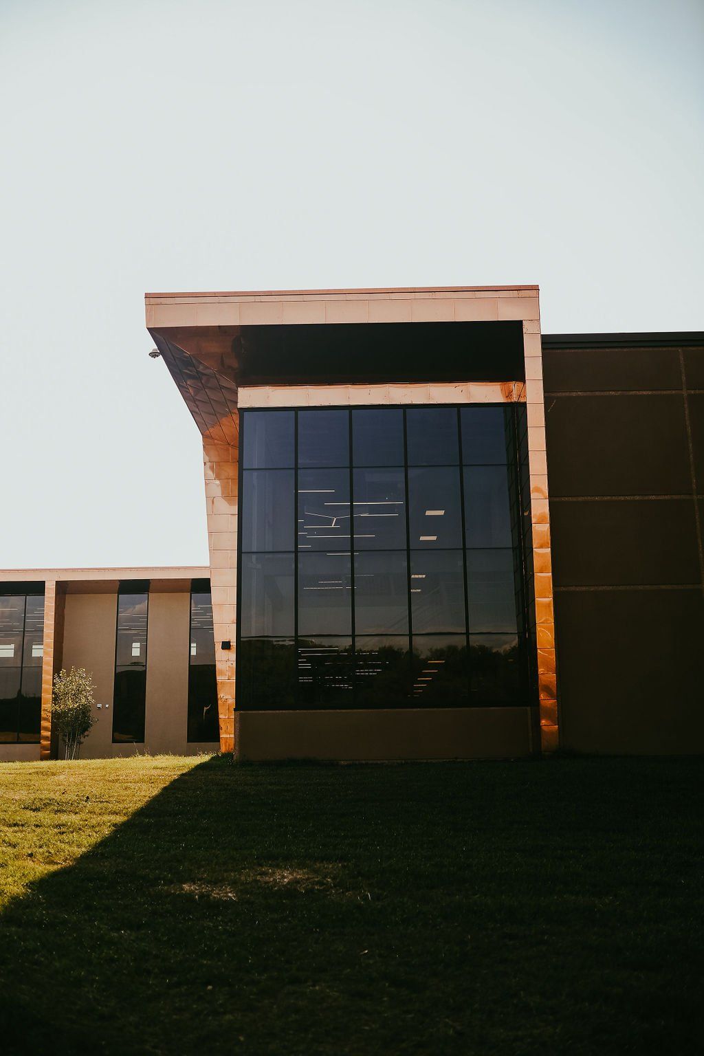 Modern building with a large glass window, a bronze-colored overhang, and grassy lawn.