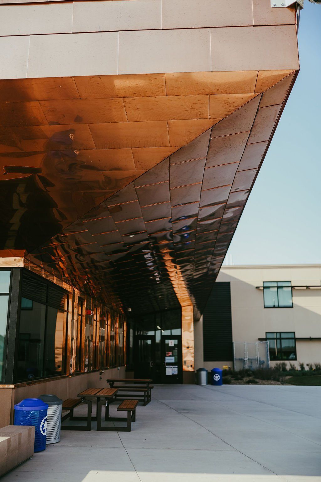 Exterior of a building with a copper-colored overhang. Tables, chairs, and windows are below. Blue sky in the background.
