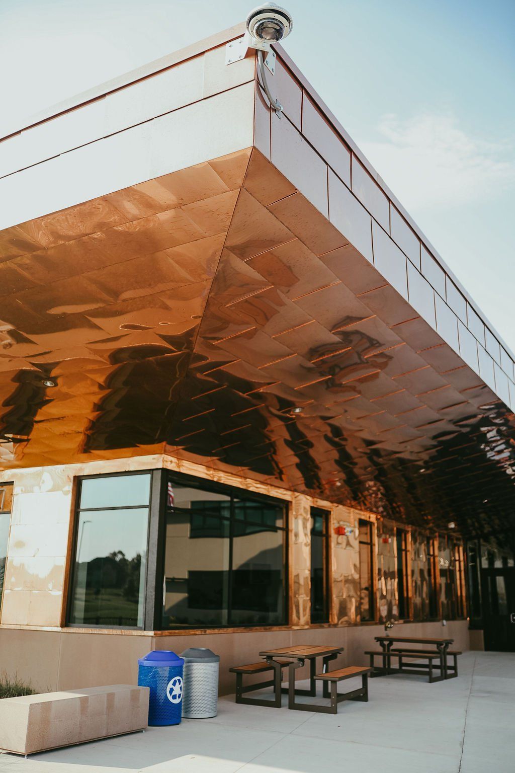 Modern building with copper-colored exterior and large windows; picnic tables and trash cans sit below.