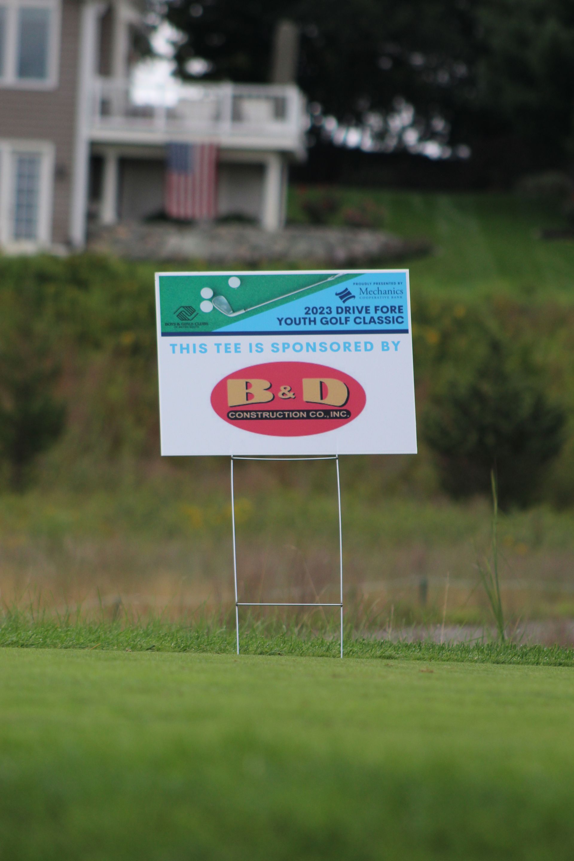 A sign is sitting on top of a lush green field in front of a house.