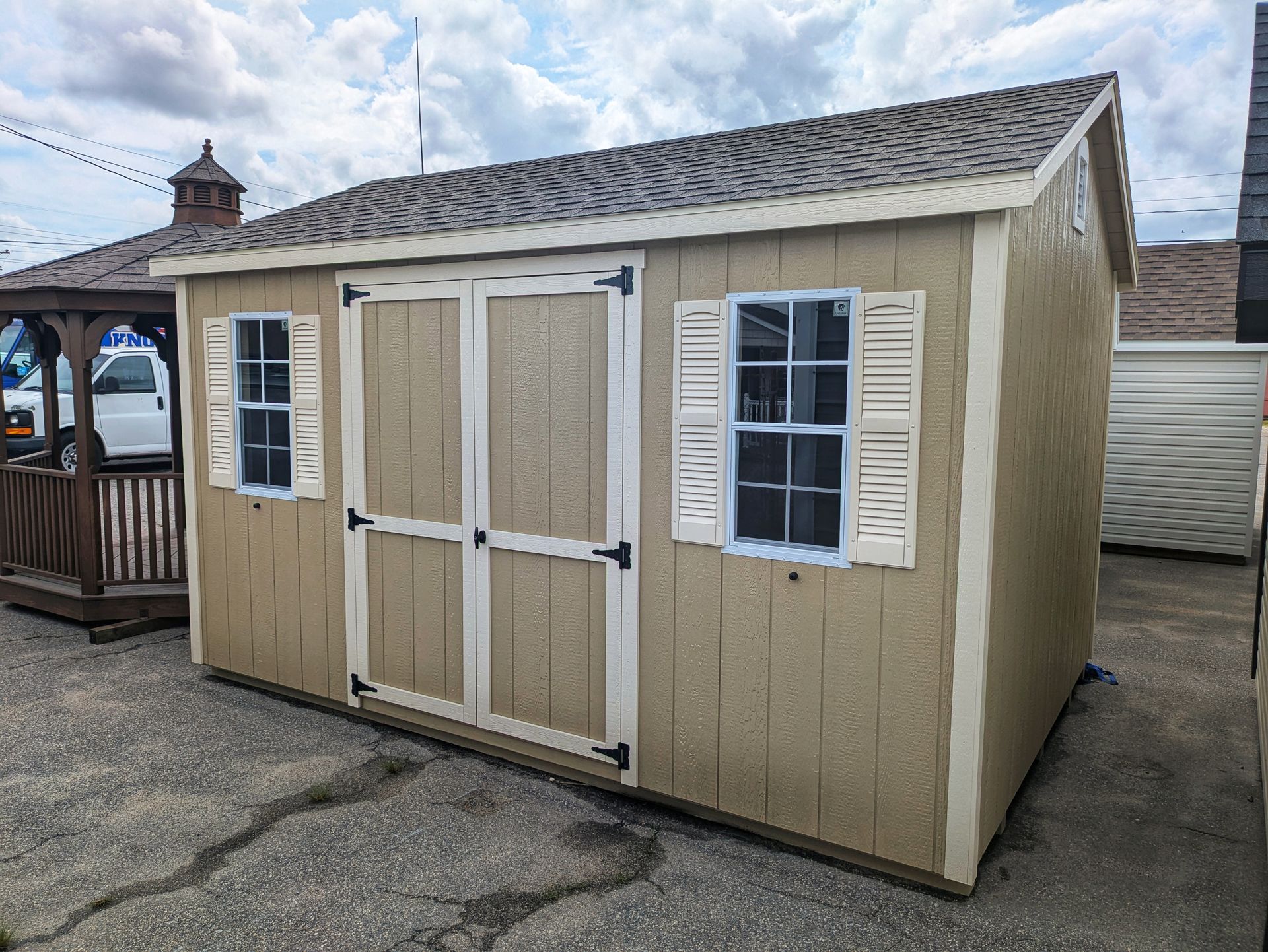 A small shed with shutters on the windows is sitting in a parking lot.
