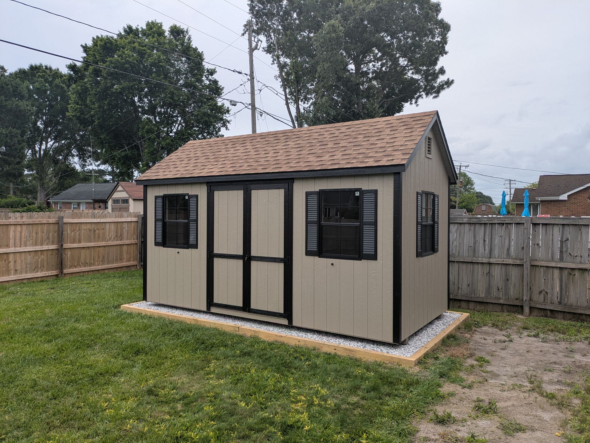 A small shed is sitting in the middle of a grassy yard next to a wooden fence.