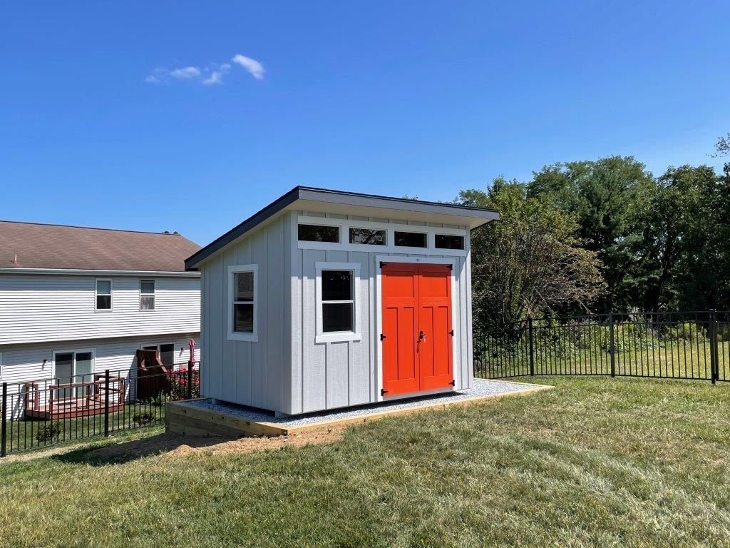 A small white shed with an orange door is sitting in the middle of a lush green field.