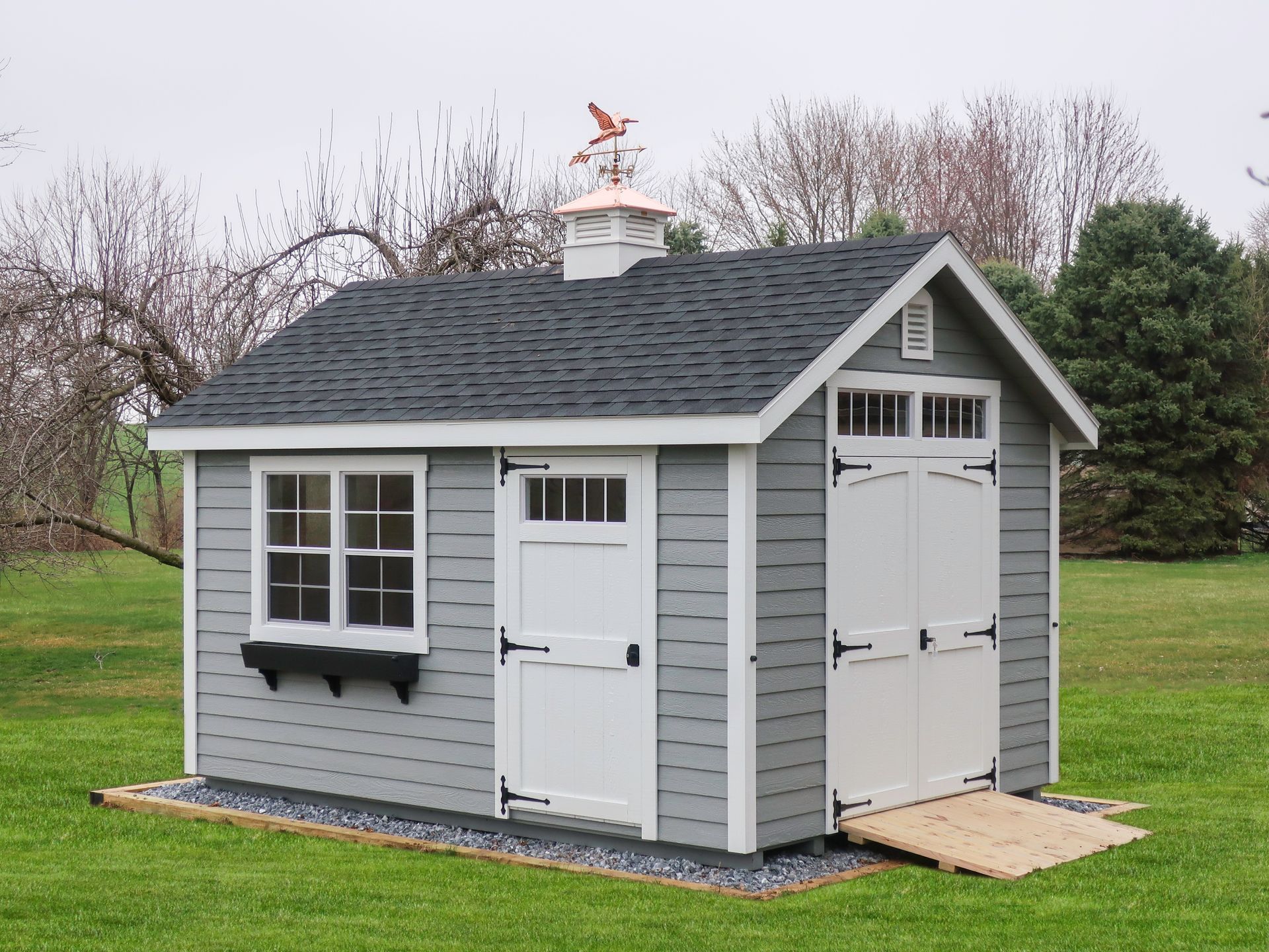 Gray shed with white trim, dark gray roof, and a copper weathervane in a grassy yard. There's a ramp on the right.