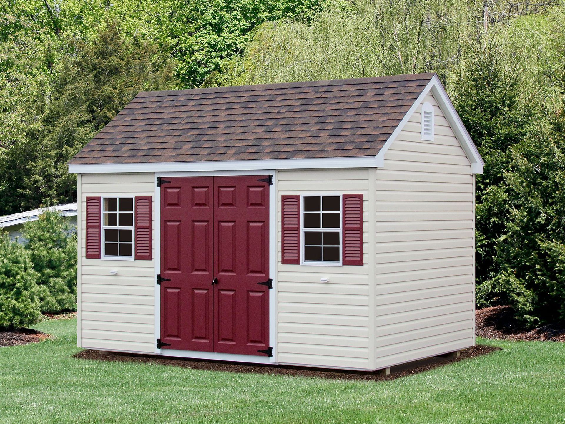 Tan shed with burgundy doors and shutters on a green lawn, surrounded by bushes.
