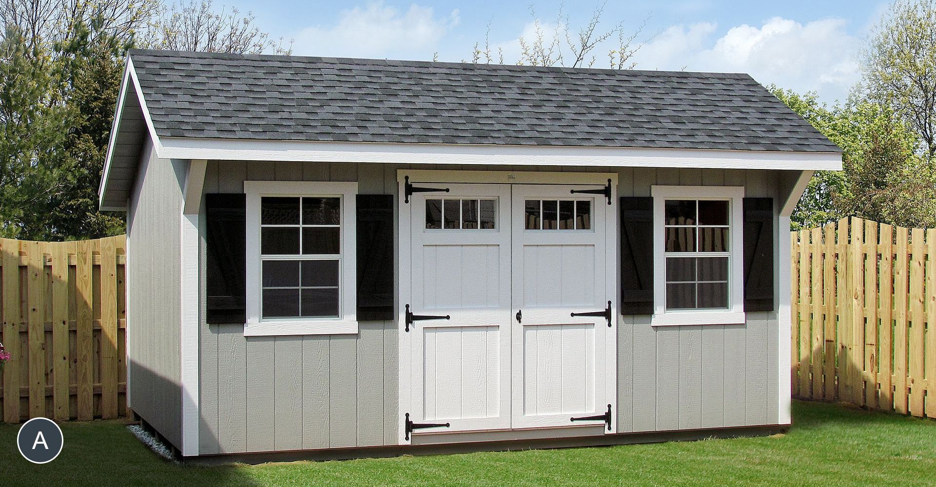 Gray shed with white doors and window frames, black shutters and a gray shingled roof, in a grassy yard next to a wooden fence.