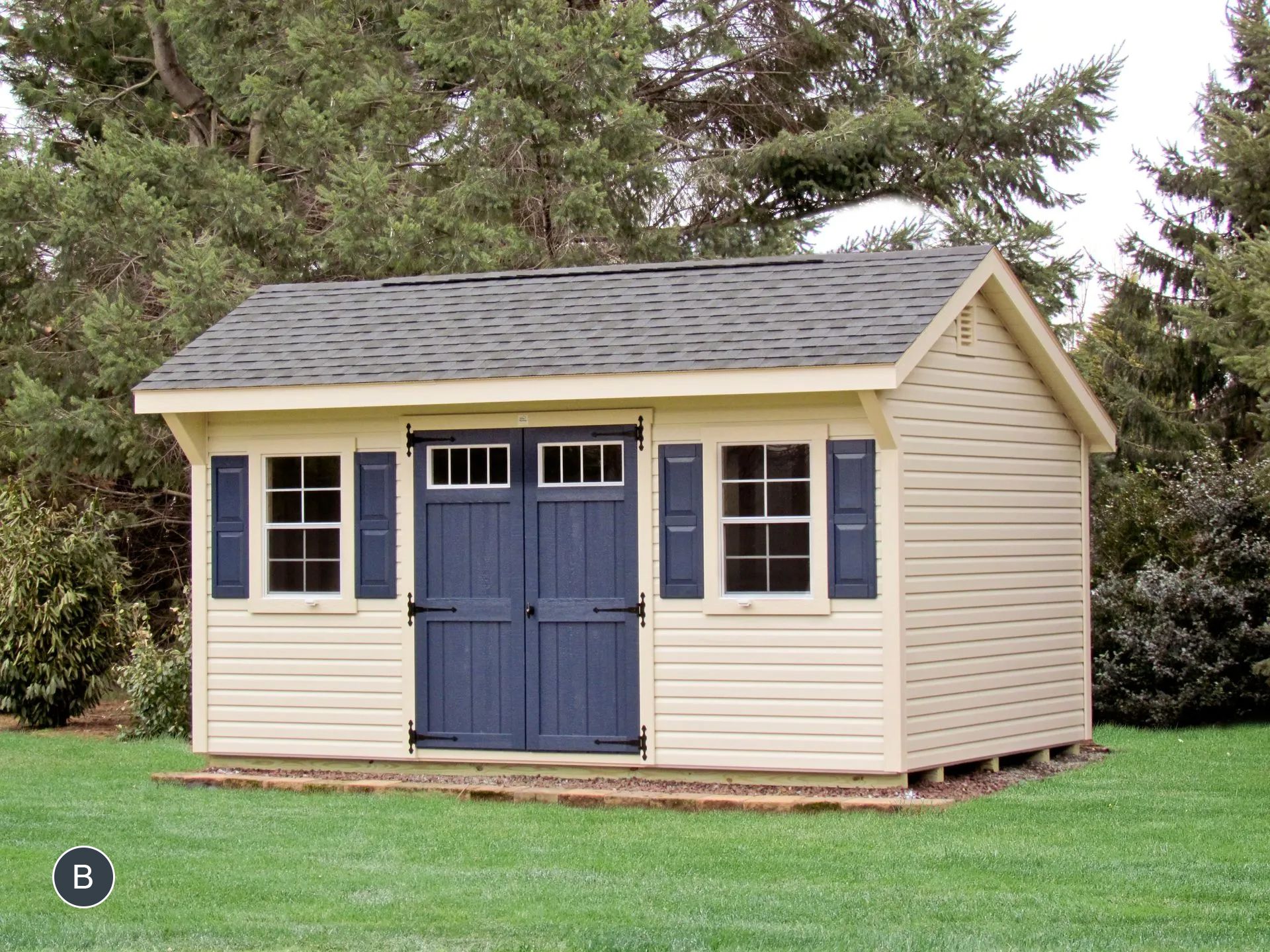 Tan shed with blue double doors and shutters, set on a green lawn surrounded by trees.