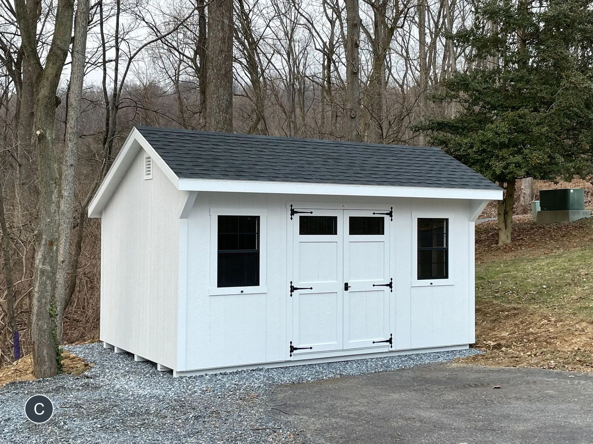 A white shed with a black roof is sitting in the middle of a gravel driveway.
