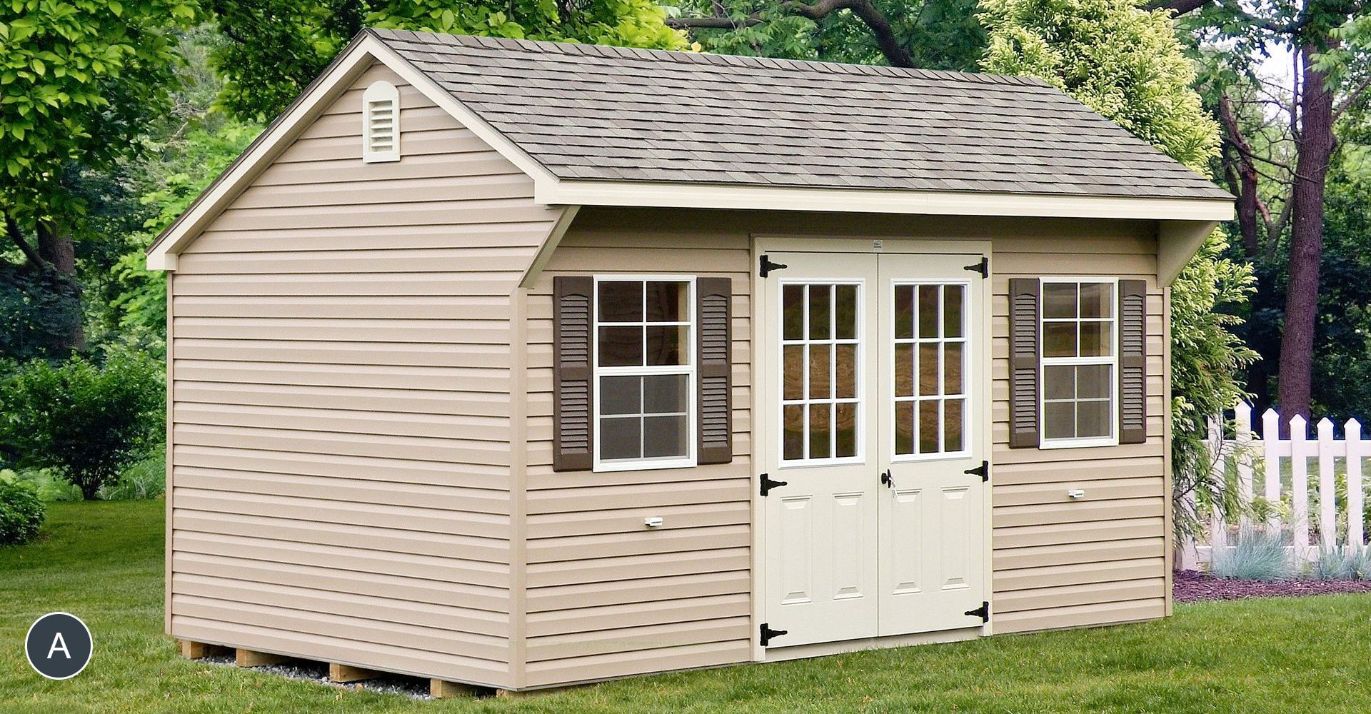 Tan shed with a gabled roof, double doors, and two windows, set in a grassy yard with a white picket fence.