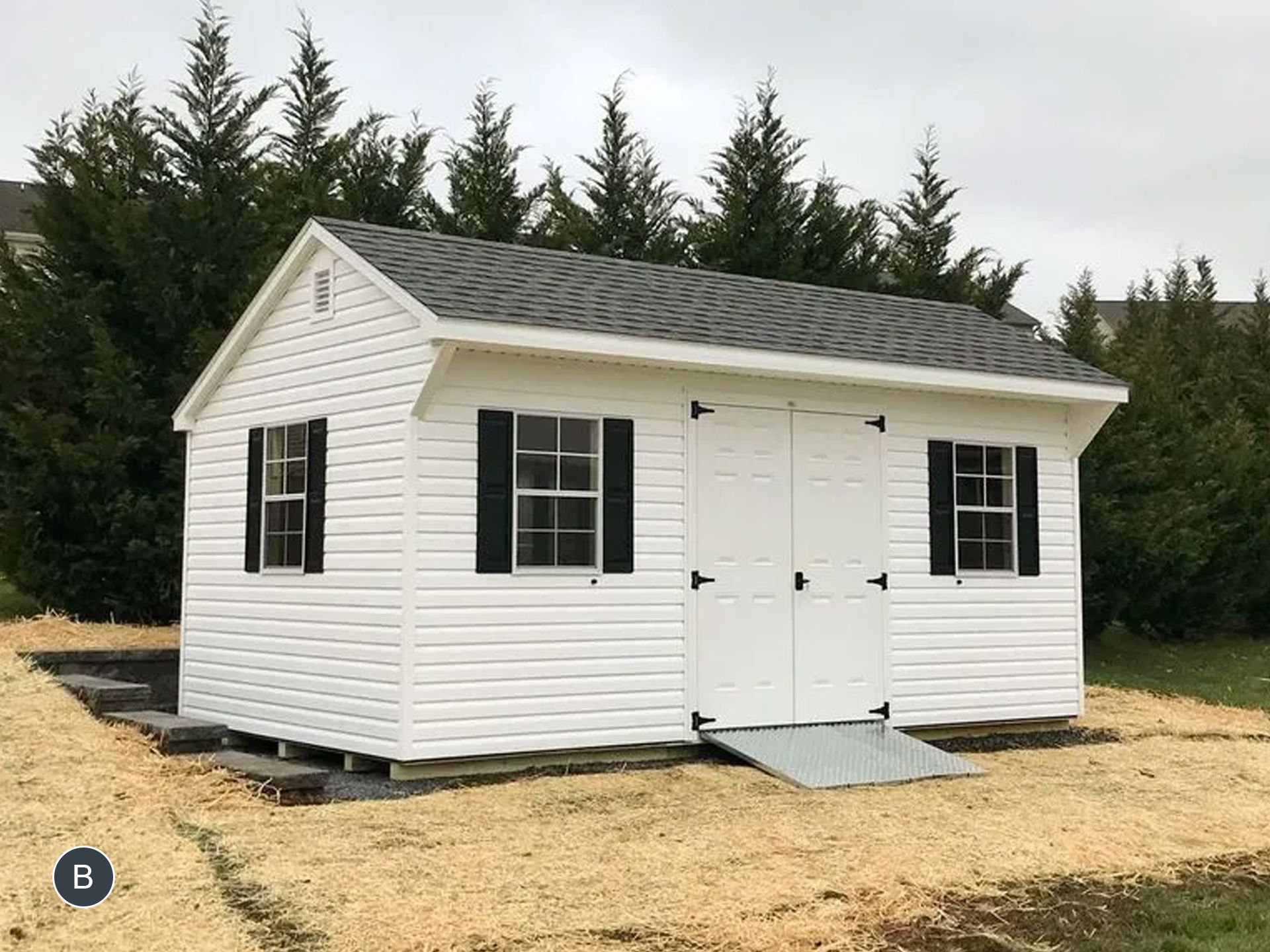 A white shed with black shutters and a ramp is sitting in the middle of a field.