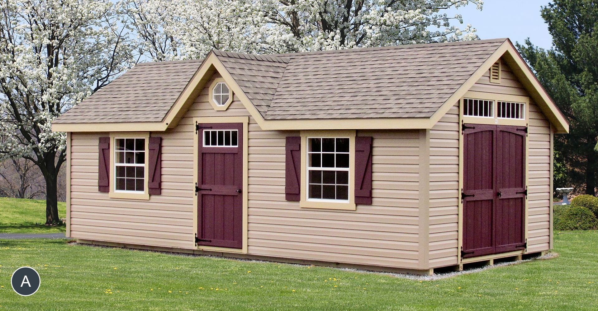 Tan storage shed with a brown roof, door, and shutters. It's in a grassy yard, under flowering trees.