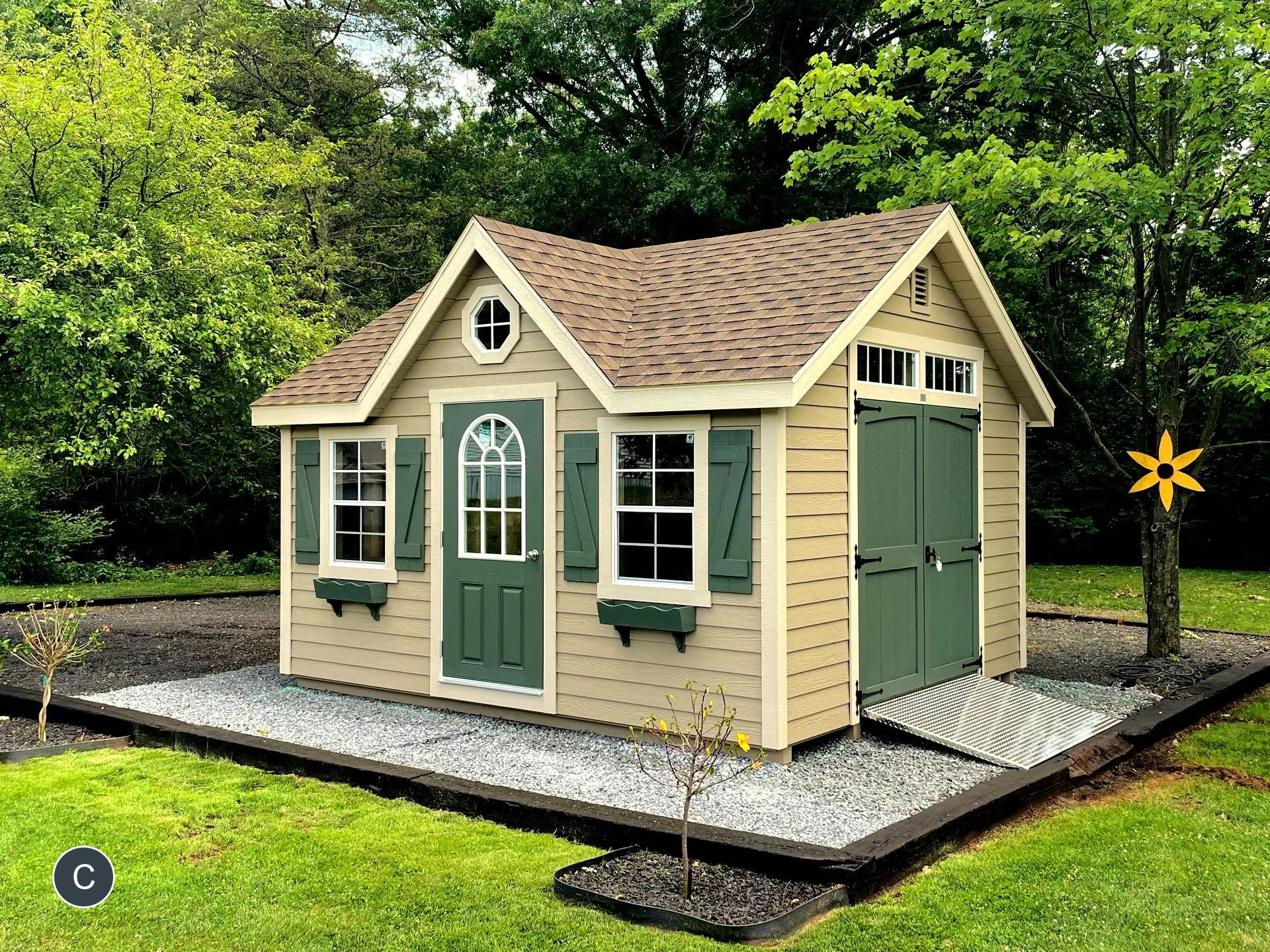 A small, tan shed with a brown roof and green door and shutters, sitting on a bed of grey stones, surrounded by grass and trees.