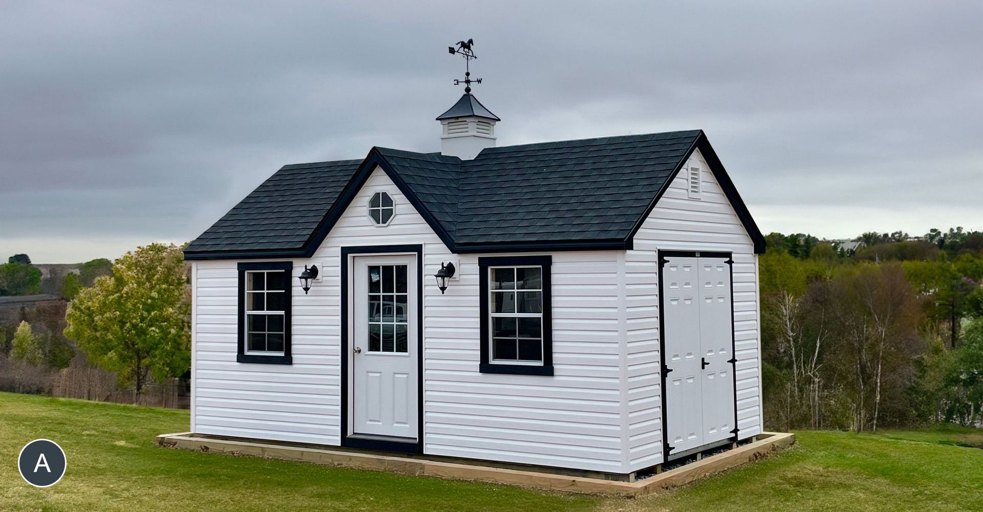 White shed with a black roof and trim, sitting on a grassy hill. Includes a decorative cupola with a weathervane.