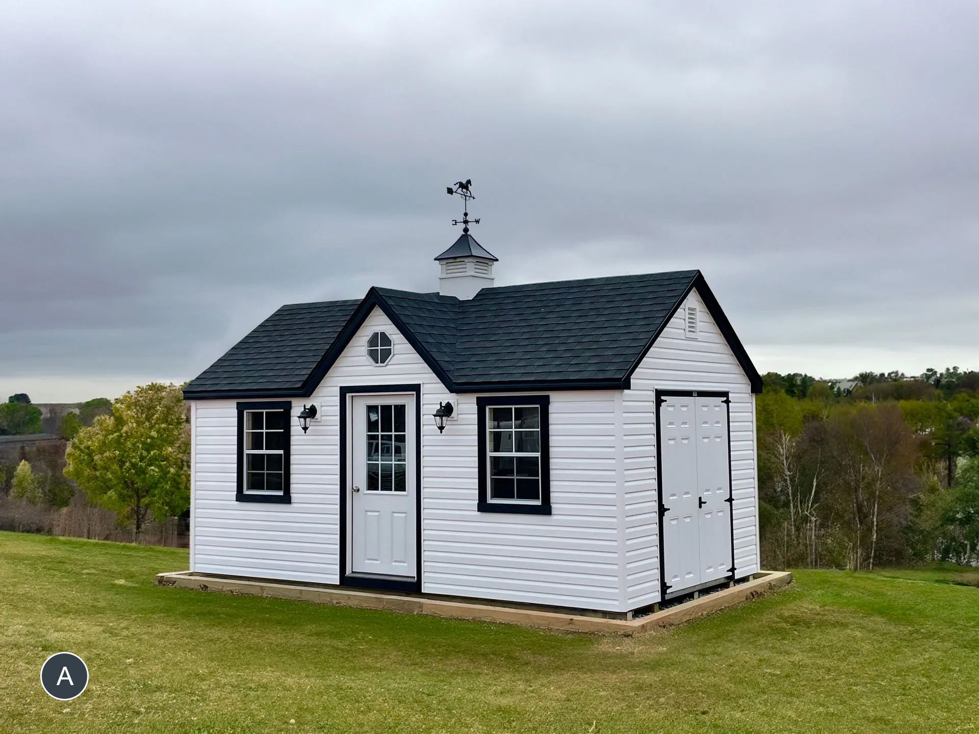 White shed with black trim and a weathervane on a grassy hill under a cloudy sky.