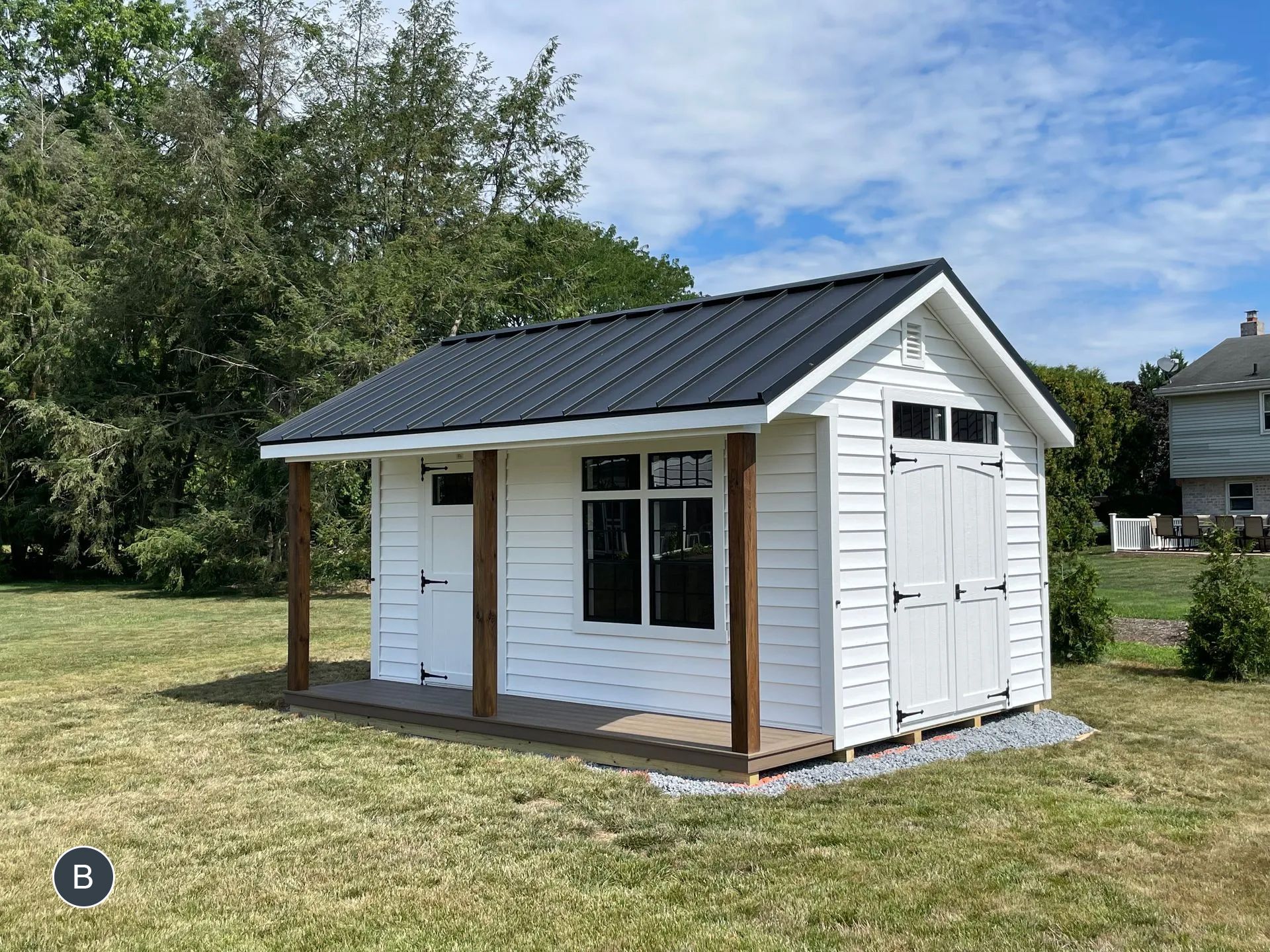 White shed with a black metal roof and wooden porch on a grassy lawn, under a blue sky.