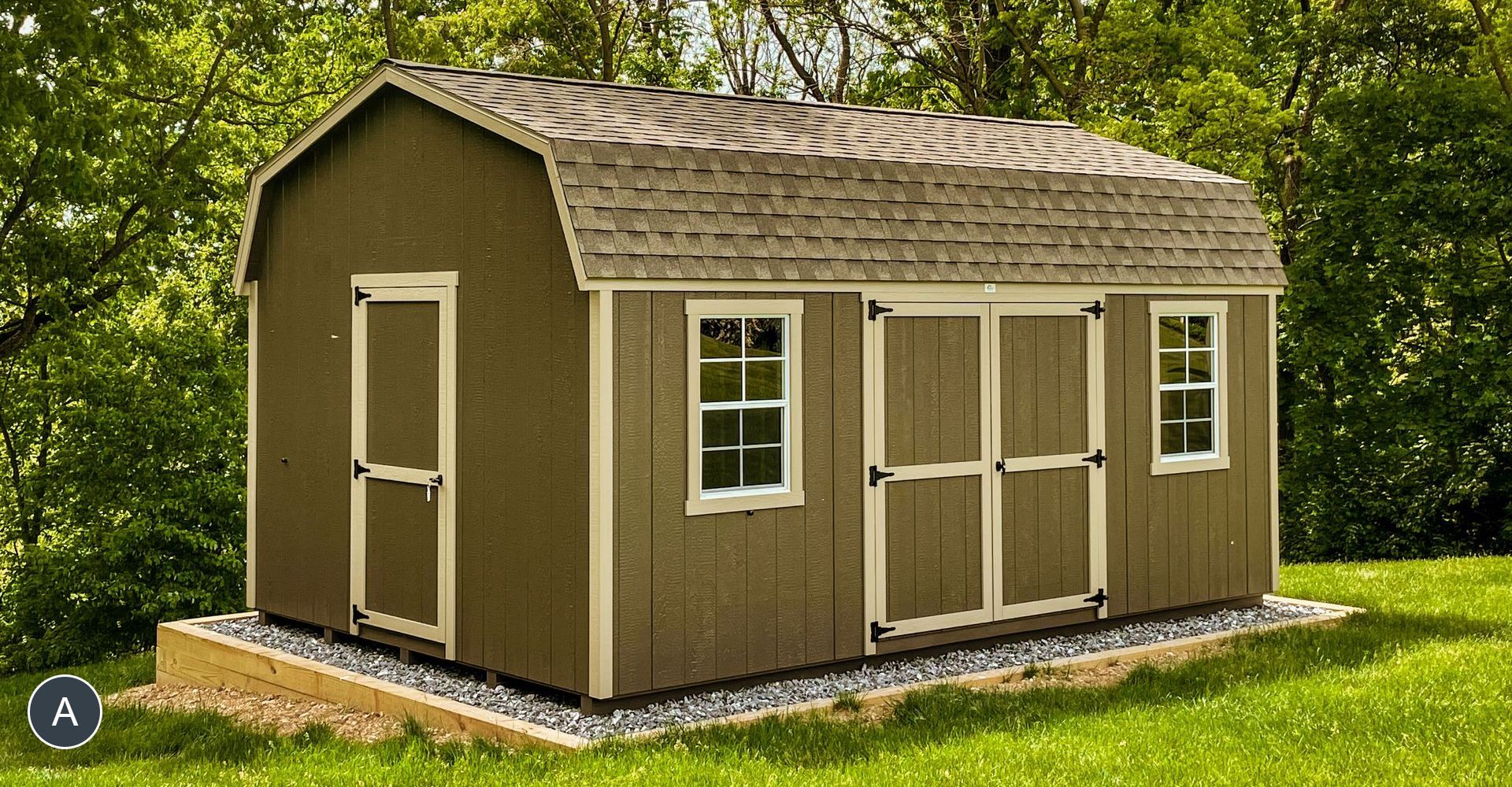 Brown shed with a barn-style roof, two windows, and two doors, set on a gravel foundation in a grassy yard.