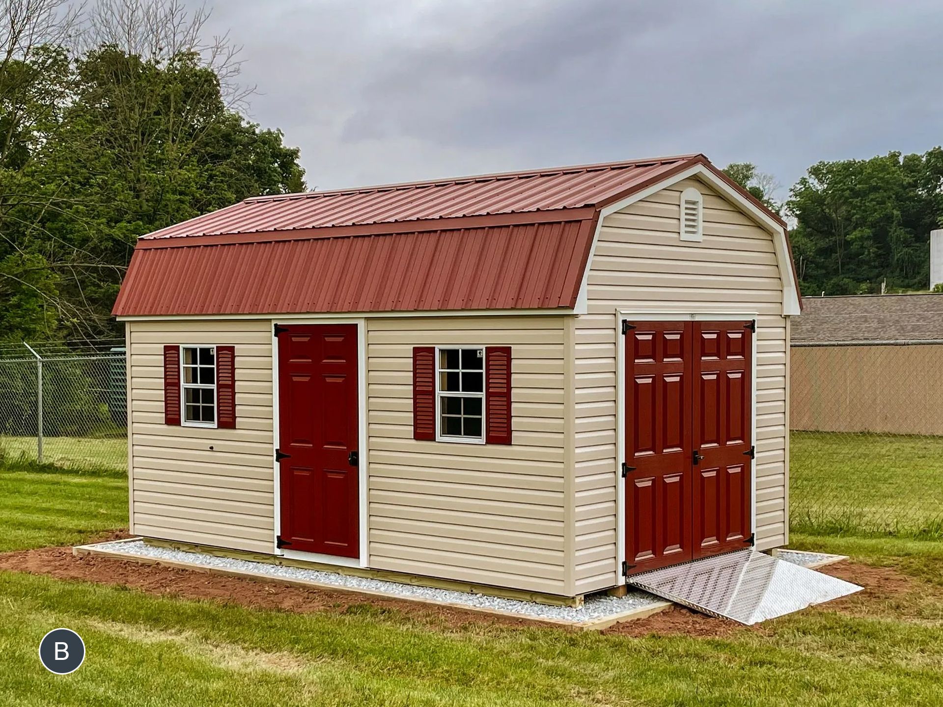 Tan shed with red doors and roof, set on a grassy yard with a ramp for the entrance.
