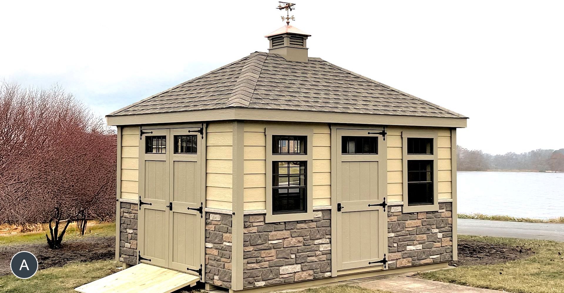 Octagonal beige and stone building with two doors and windows, topped with a weather vane. It's near water.
