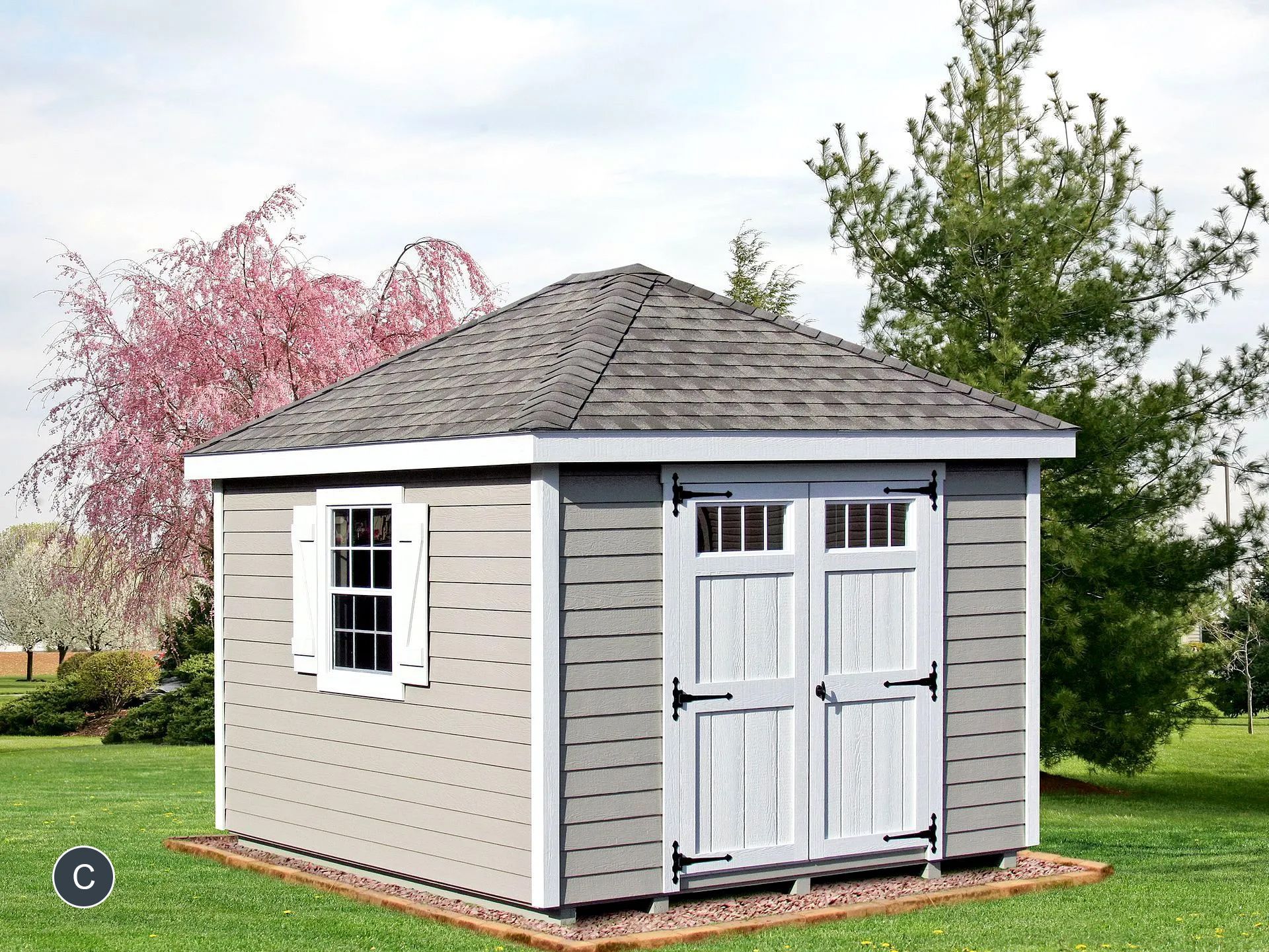 Gray shed with a gray shingle roof, white doors, and window on a grassy lawn.