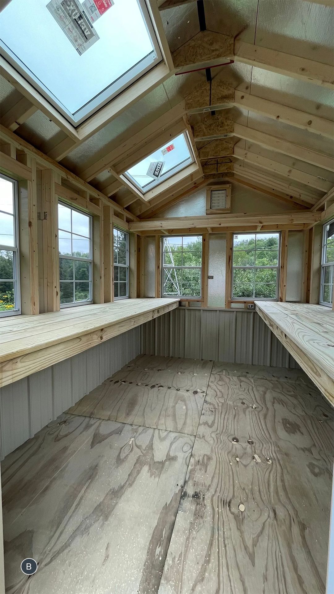 Interior of a wood-framed greenhouse under construction, featuring multiple windows, skylights, and work surfaces.