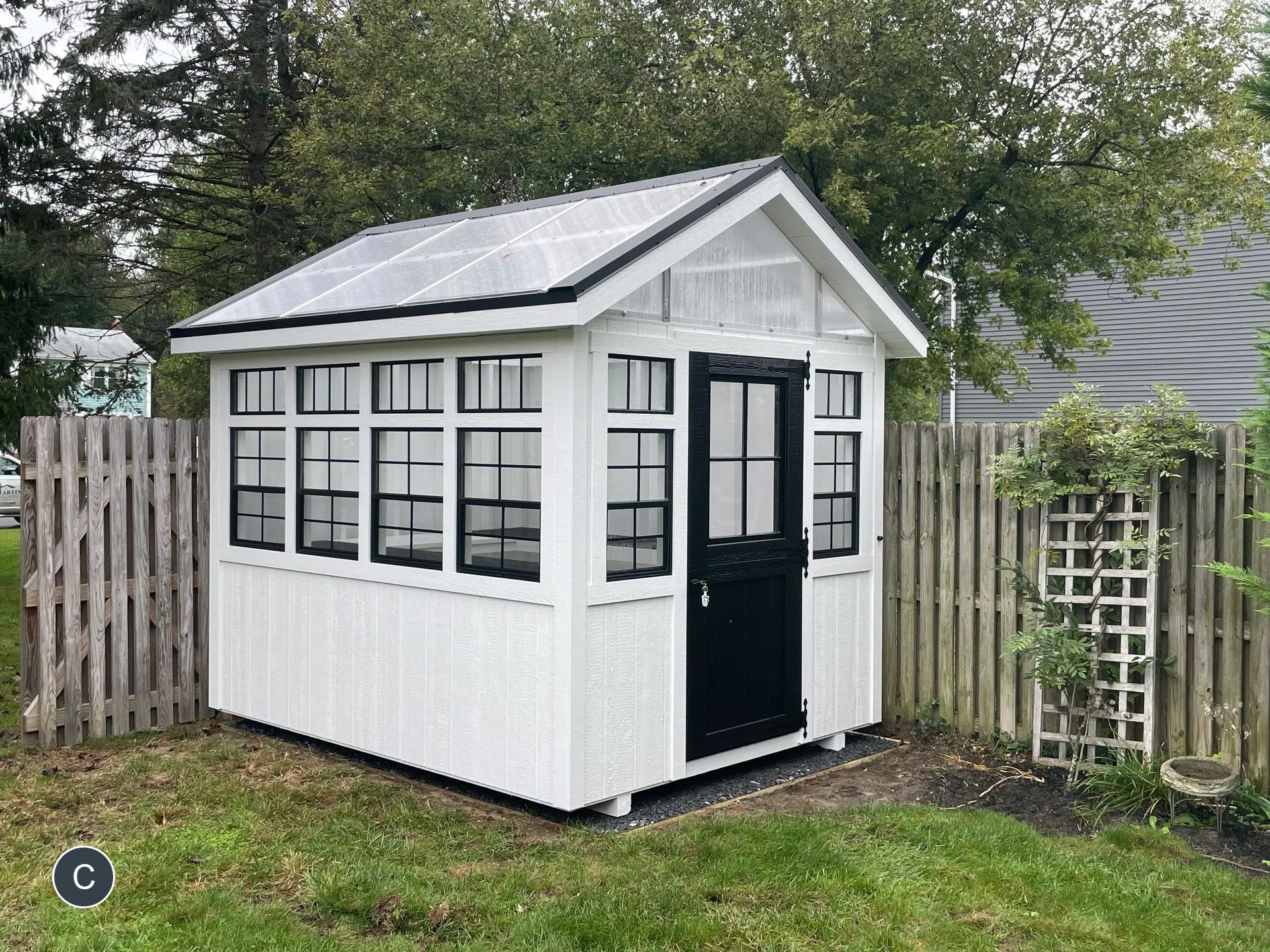 White and black garden shed with a transparent roof, set between wooden fences. Green grass and trees surround it.