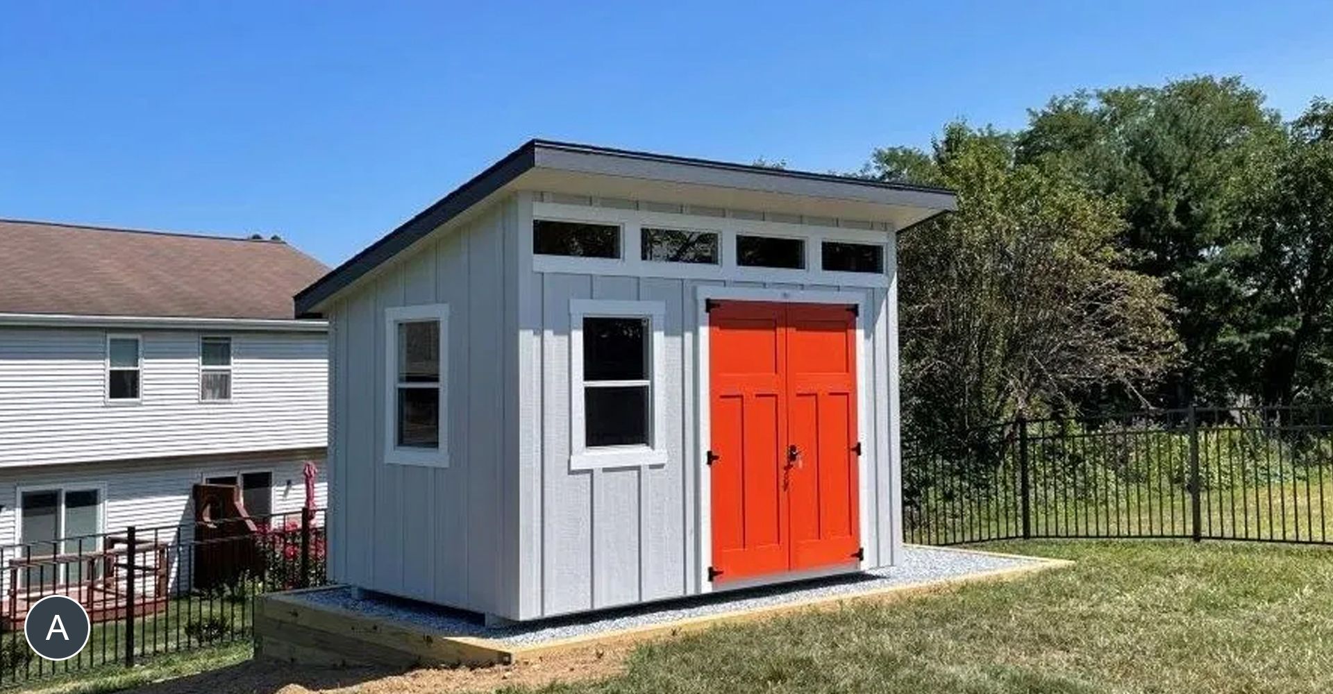 A small, modern shed with gray siding and a bright orange door stands in a grassy yard on a sunny day.