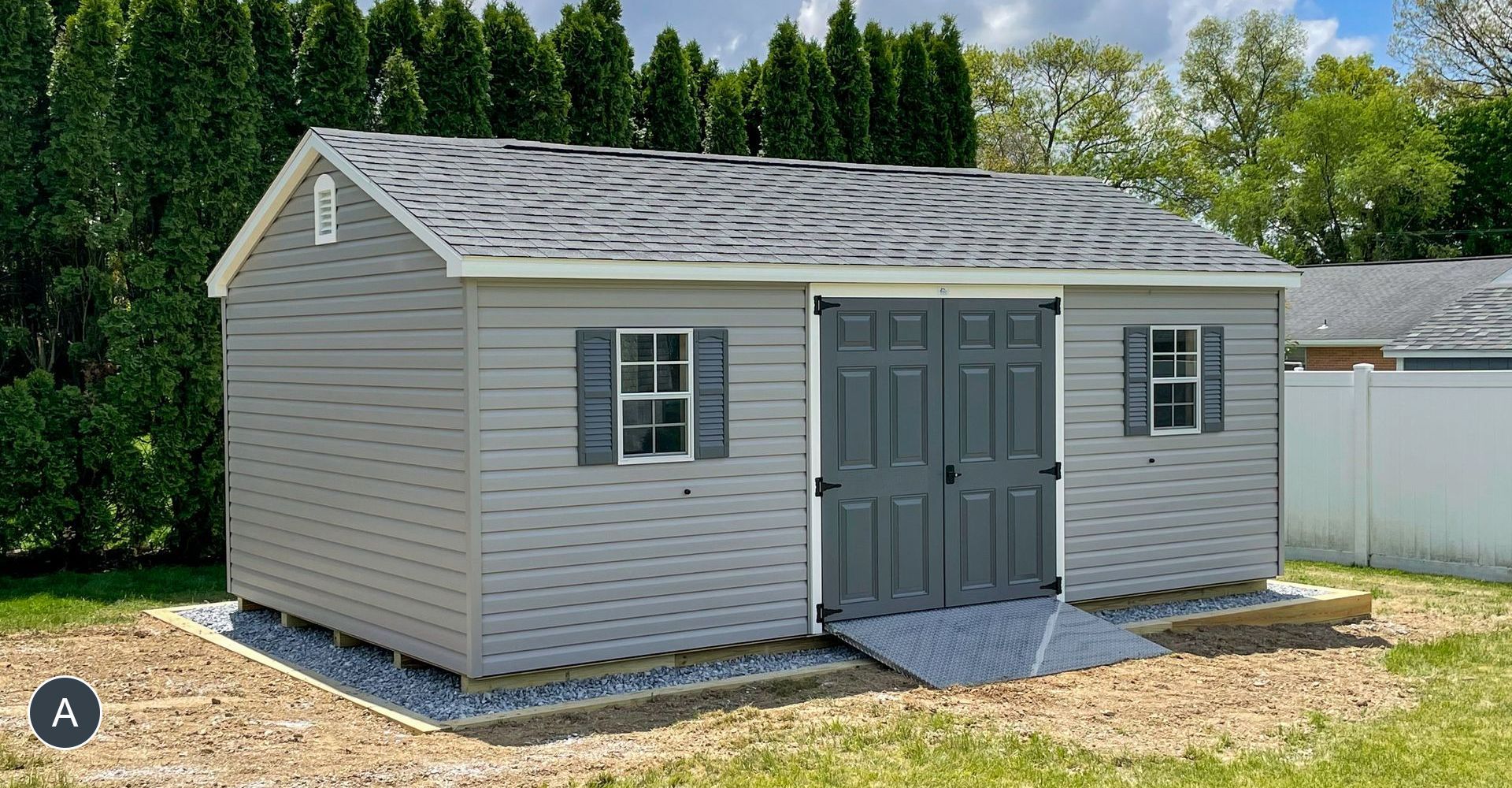 Gray shed with double doors and a ramp, two small windows with shutters, and a gravel foundation.