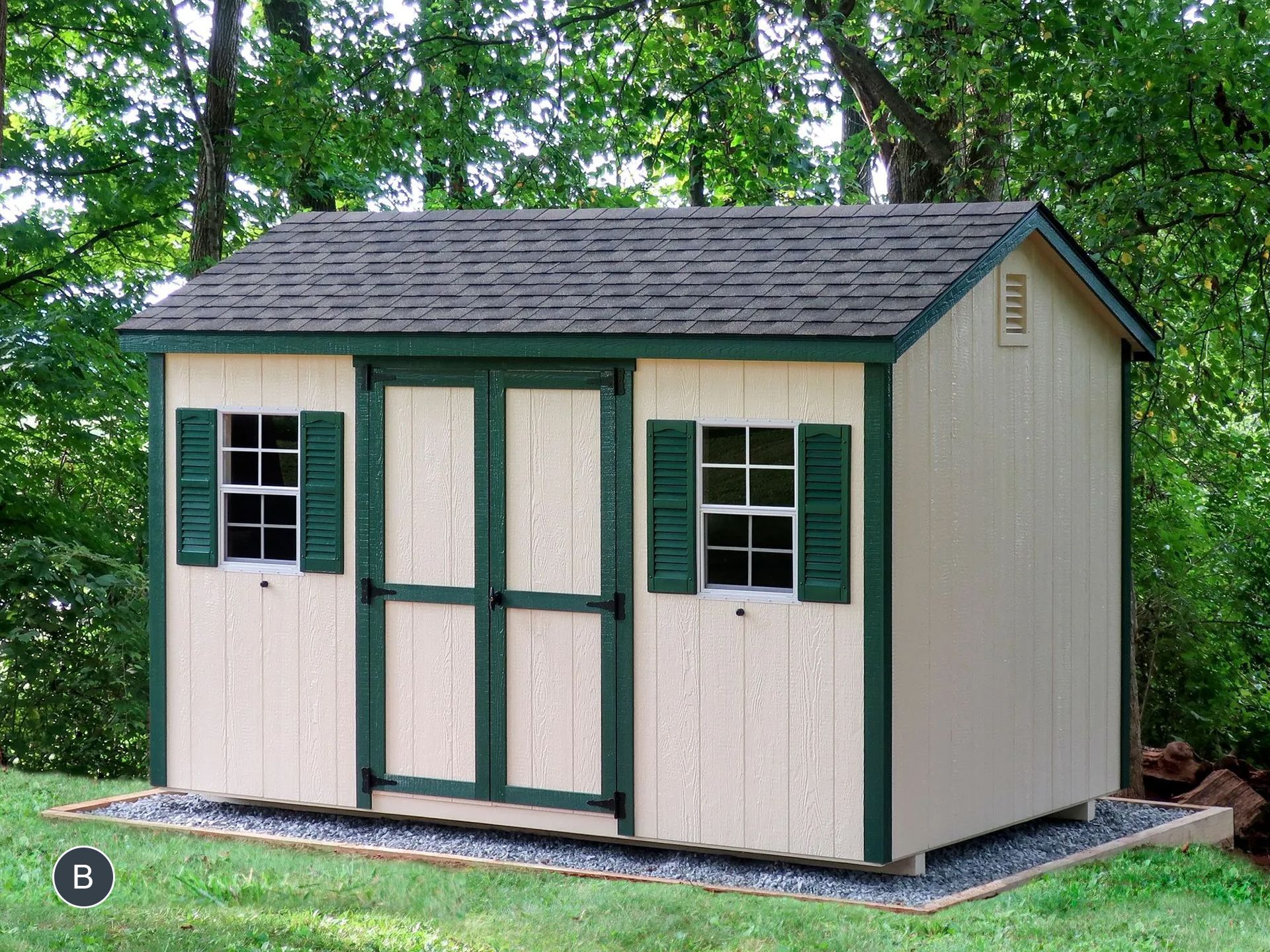 Tan and green shed with dark green shutters and door, on a gravel base in a grassy yard surrounded by trees.