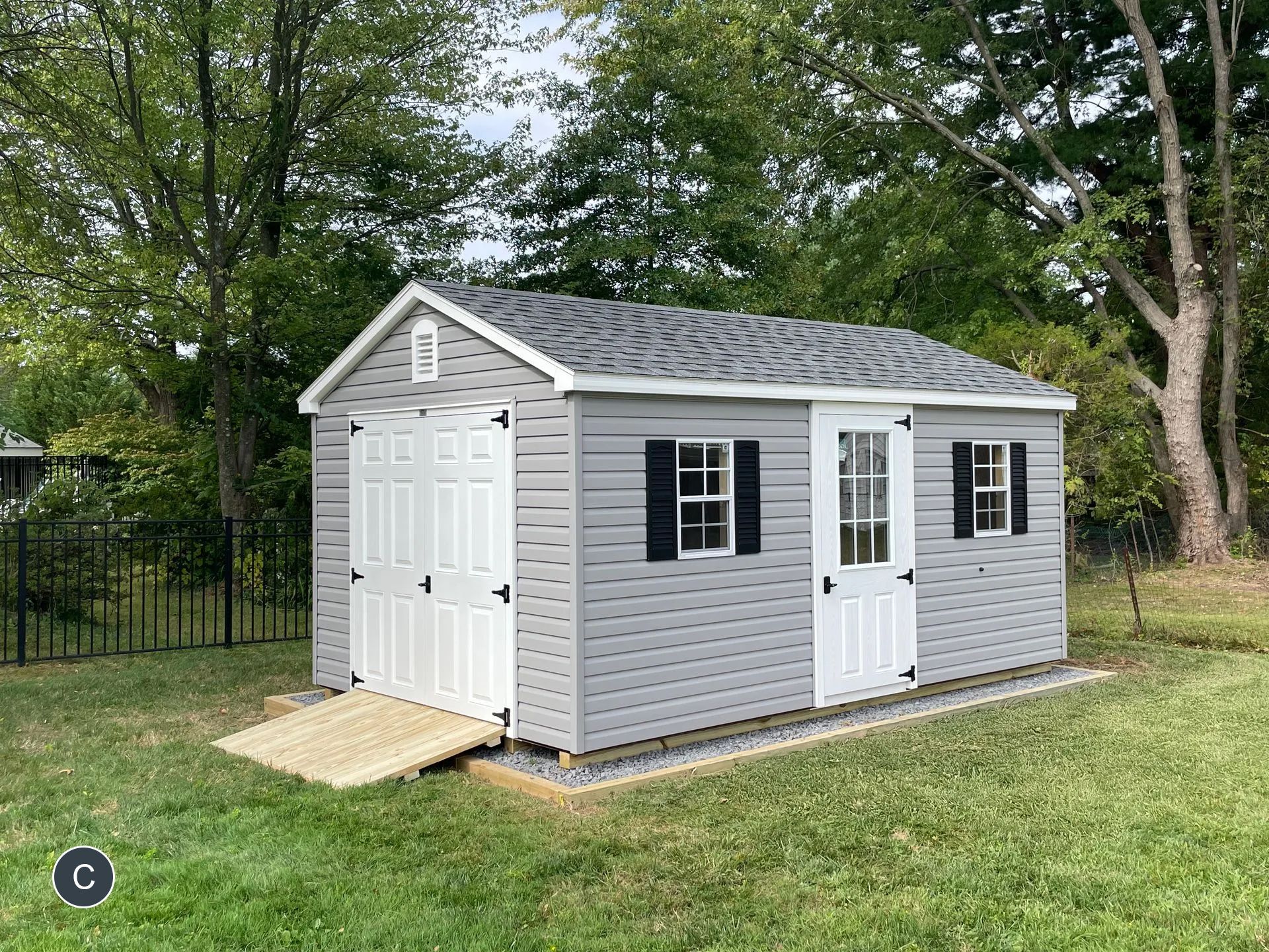 Gray shed with white doors and ramp in a grassy backyard; black shutters and dark roof.