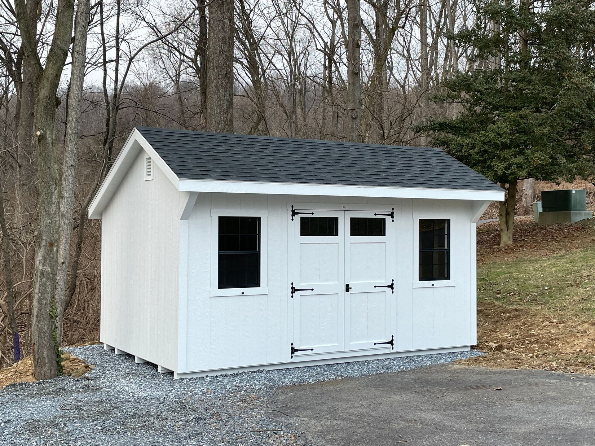A white shed with a black roof is sitting in the middle of a gravel driveway.