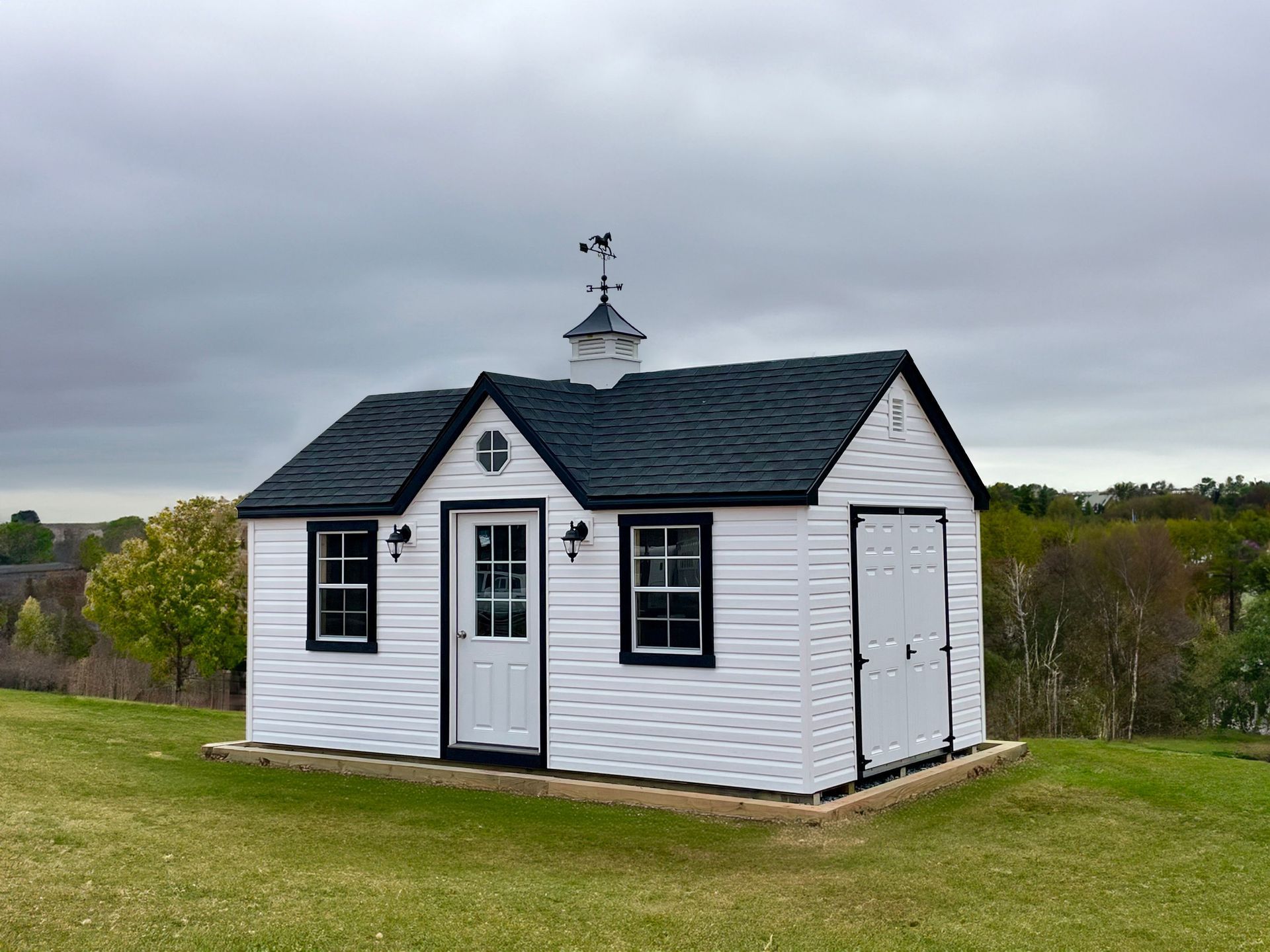A small white shed with a black roof is sitting in the middle of a grassy field.