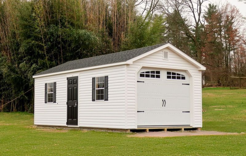 A white garage with a black door is sitting in the middle of a grassy field.