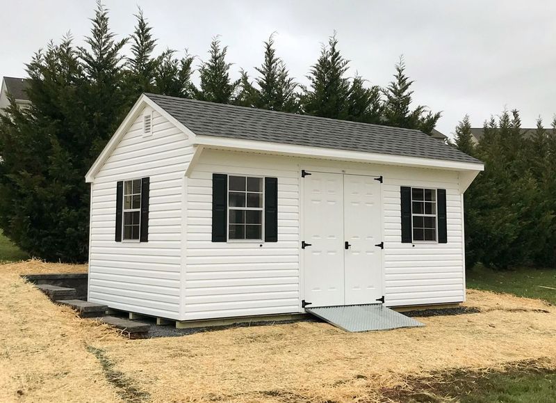 A white shed with black shutters and a ramp is sitting in the middle of a field.