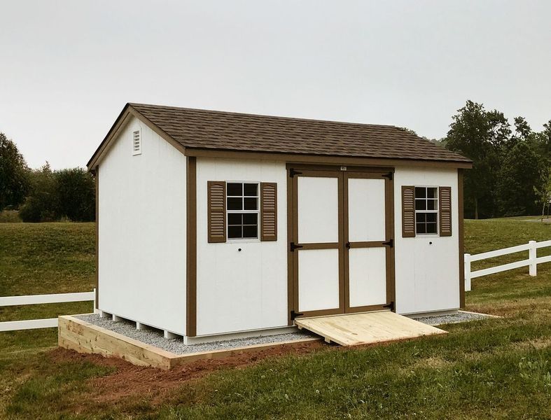 A white shed with brown shutters is sitting in the middle of a grassy field.