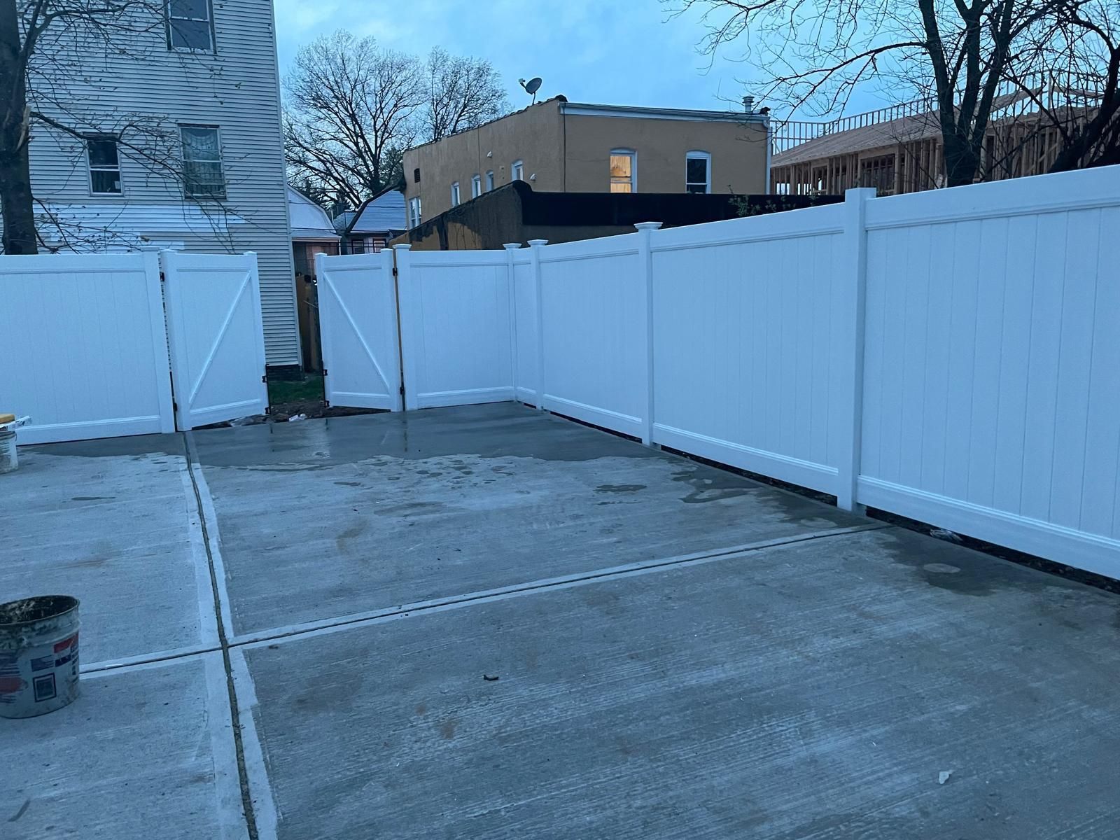 A white fence surrounds a concrete driveway in a backyard.